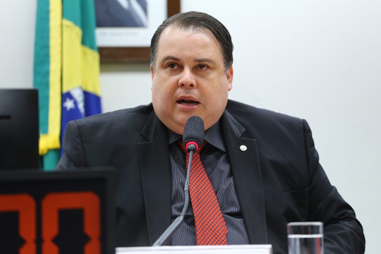 Deputado Julio Cesar Ribeiro fala ao microfone. Ele é branco e tem o cabelo escuro, usa um terno preto com gravata vermelha