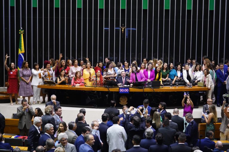 Deputadas estão em pé atrás de uma grande mesa de madeira no Plenário da Câmara