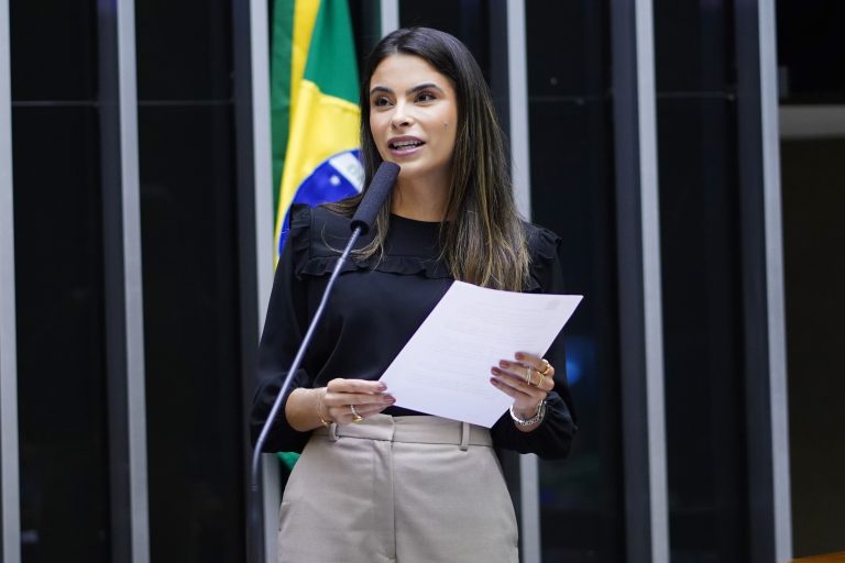 Deputada Yandra Moura fala ao microfone. Ela é uma mulher branca, com cabelos escuros e longos, uma uma calça bege e blusa preta e segura um papel