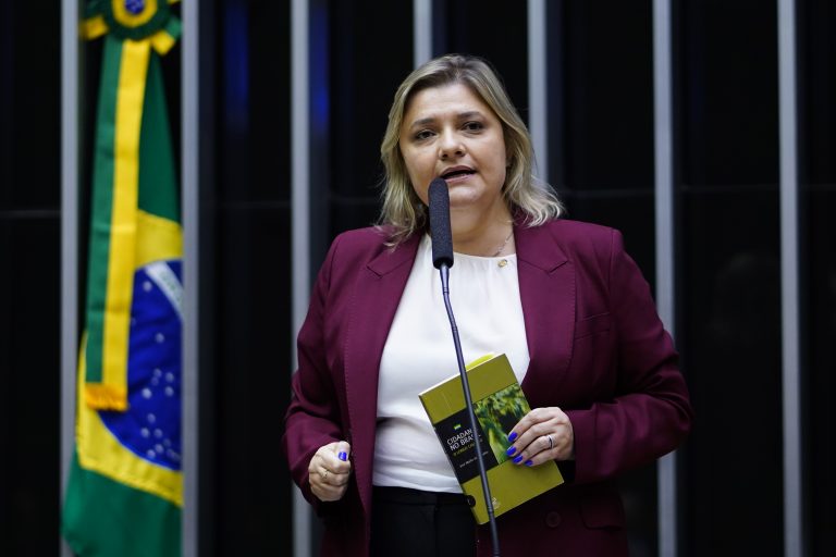 Deputada Professora Luciene Cavalcante fala ao microfone. Ela é branca, tem o cabelo longo e loiro, usa uma blusa branca e um blazer vinho e segura um livro