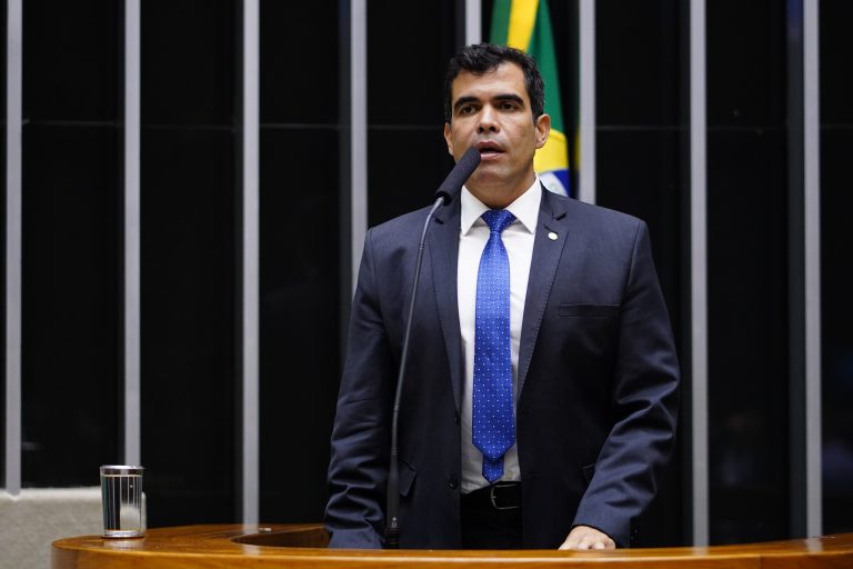 Deputado Ricardo Ayres fala ao microfone. Ele é claro, tem cabelo escuro e veste um terno azul-marinho