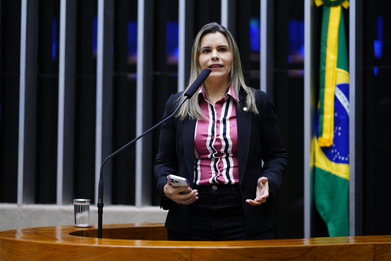 Deputada Clarissa Tércio fala ao microfone. Ela é branca, tem o cabelo loiro, usa um terno preto e blusa listrada de rosa, preto e branco, e segura um celular