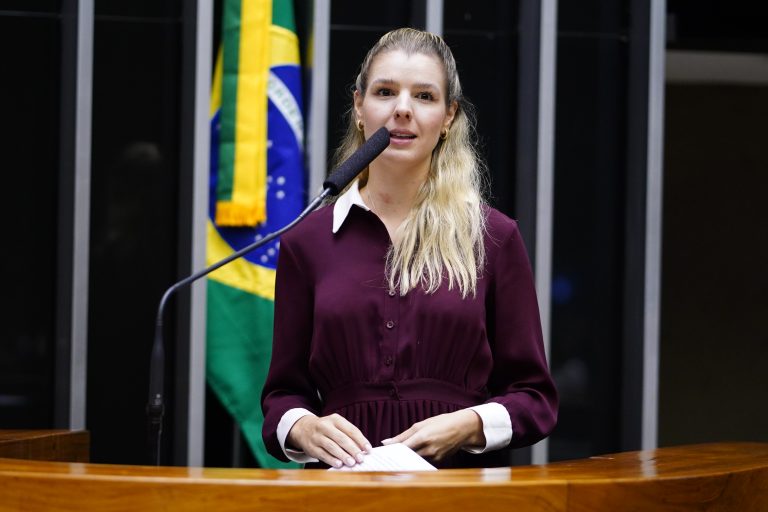 Deputada Luísa Canziani fala ao microfone. Ela é branca, loira e usa um vestido vinho