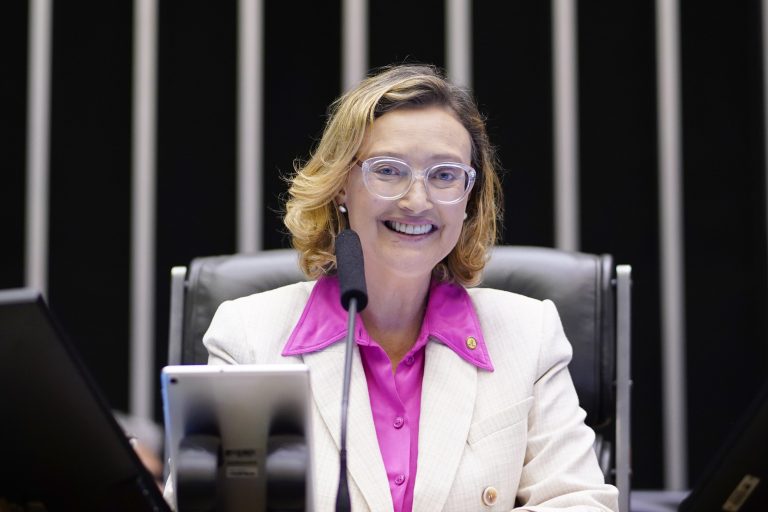 Deputada Maria do Rosário está sentada. Ela é branca, tem o cabelo médio e loiro, usa óculos, uma camisa rosa e um blazer claro. Ela olha para câmera e sorri.