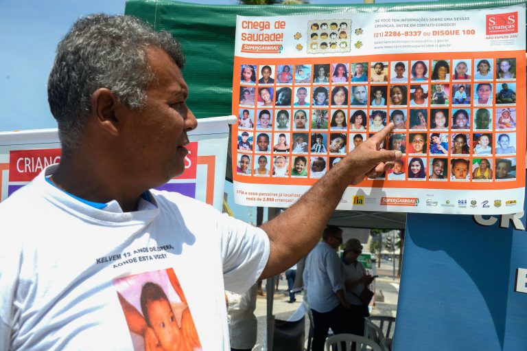 Foto de um pai apontando a imagem de uma criança num cartaz cheio de fotos de crianças desaparecidas