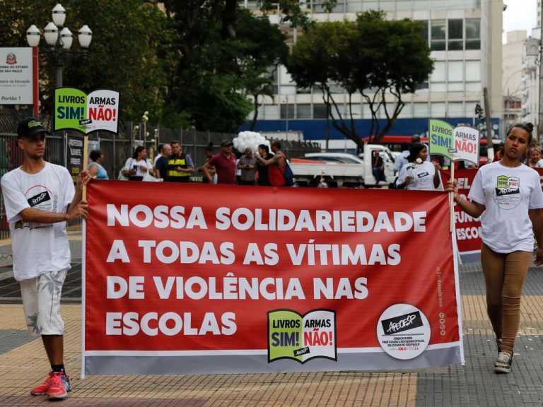 Pessoas protestam contra a violência nas escolas. Elas estão reunidas numa rua e seguram cartazes