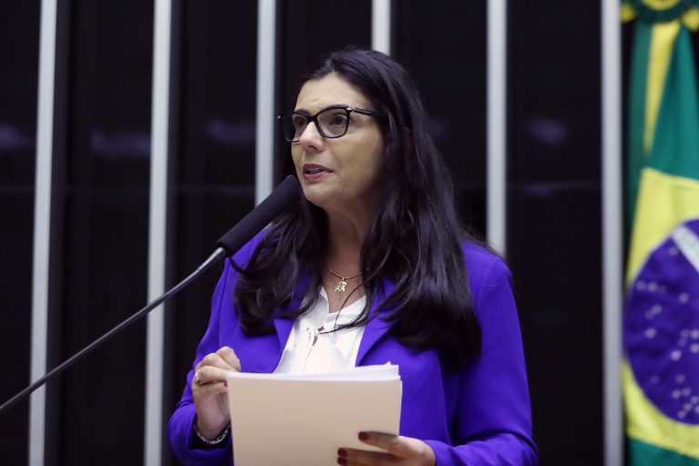 Deputada Delegada Ione fala ao microfone. Ele á branca, tem o cabelo longo e escuro, usa óculos e um blazer azul e segura um papel