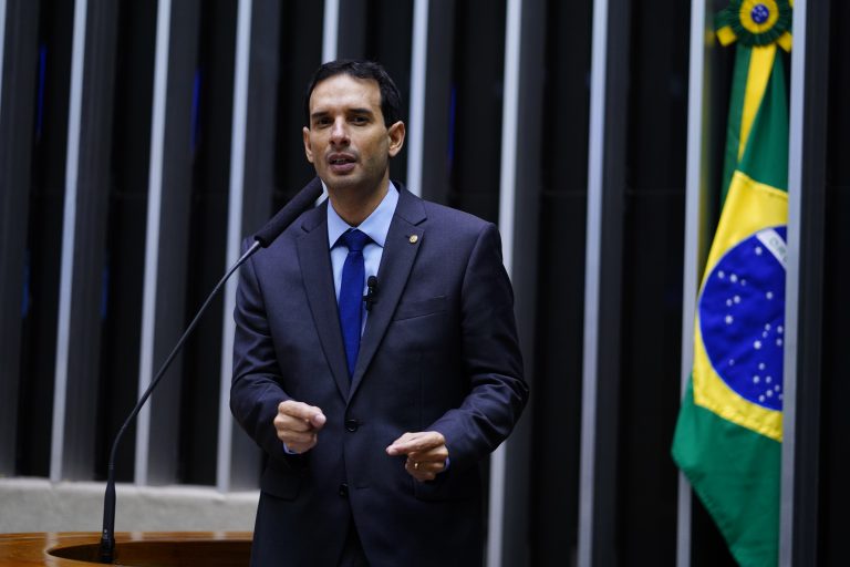 Deputado Leo Prates fala ao microfone. Ele é claro, tem o cabelo escuro, usa um terno azul-marinho e atrás dele, no lado direito, tem uma bandeira do Brasil