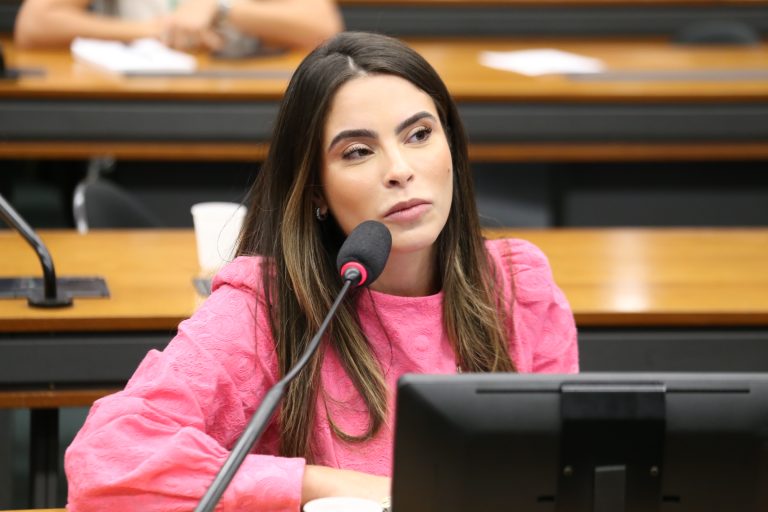 Deputada Yandra Moura fala ao microfone. Ela é branca, tem o cabelo longo e escuro e usa uma blusa rosa