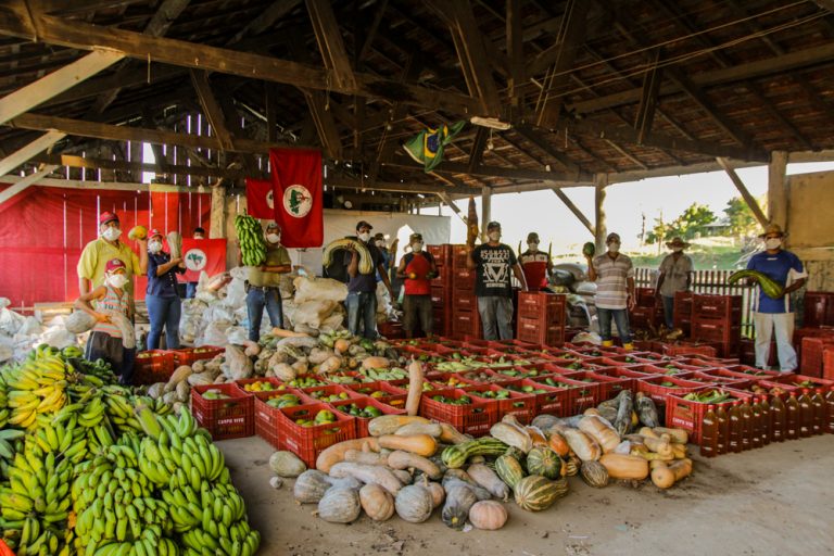 Frutas e verduras em caixas. Ao fundo pessoas em pé e uma bandeira do MST estendida