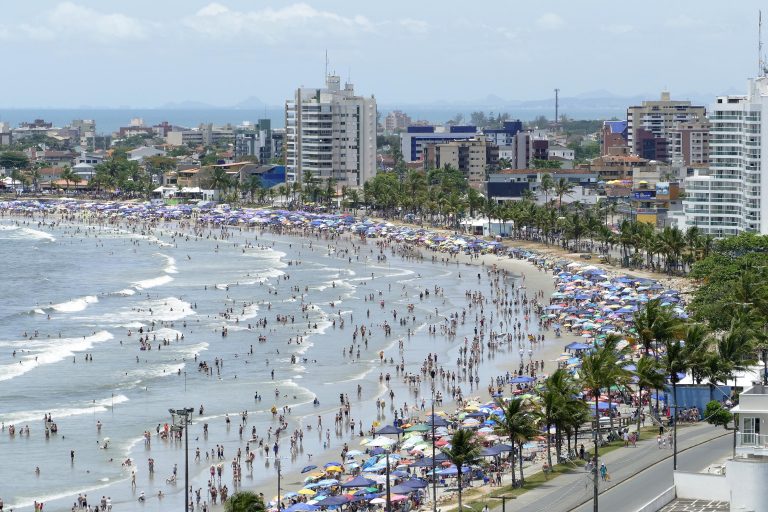 Vista de uma praia lotada num dia de sol. Ao fundo prédios altos