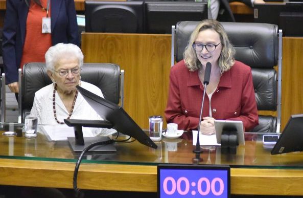 Duas mulheres estão sentadas à mesa, enquanto uma delas fala ao microfone