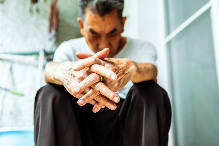 Imagem desfocada de um homem sentado, encostado na parede, com as mãos cruzadas em cima dos joelhos e a cabeça baixa