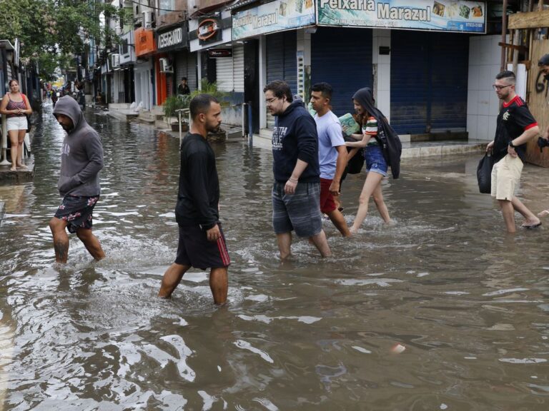 Diversas pessoas atravessam a pé uma rua inundada pelas chuvas. A água bate na canela das pessoas