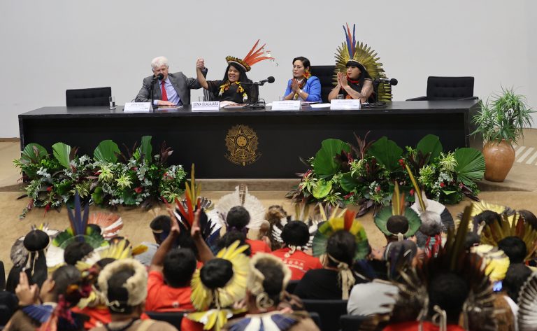 Vários índios usam cocar e estão sentados num plenário