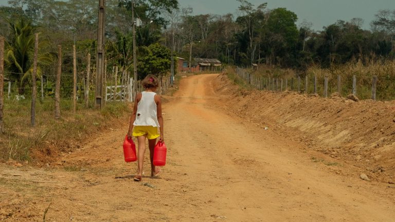 Uma menina segura dois garrafões de água e caminha numa estrada de terra vermelha