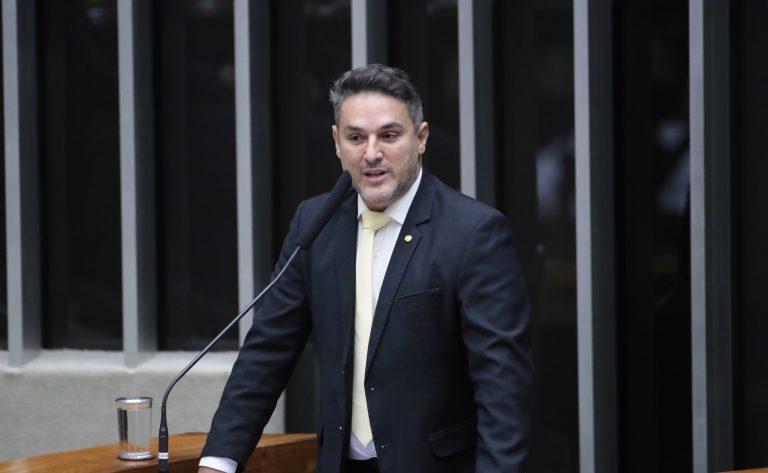 Deputado Zucco fala ao microfone. Ele é um homem claro, com cabelo curto e escuro. Veste um terno azul-marinho e segura um papel