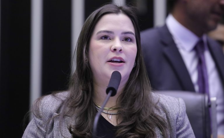 Deputada Maria Arraes fala ao microfone. Ela é clara, tem cabelos escuros, lisos e compridos; e usa um blazer cinza