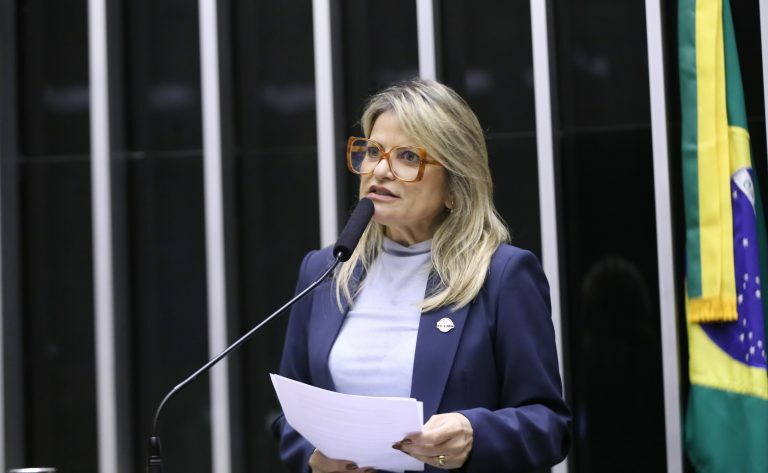 Deputada Flávia Morais fala ao microfone. Ela é loira, tem o cabelo comprido, usa uma blusa branca e segura um papel