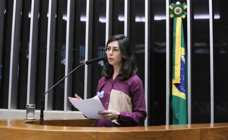 Uma mulher branca discursa. Ela tem cabelo escuro e veste uma camisa rosa