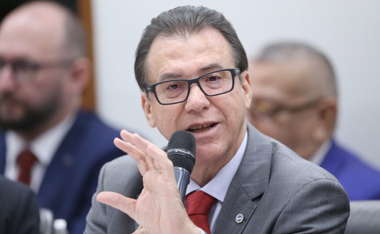Ministro Luiz Marinho fala ao microfone. Ele é um homem claro, tem cabelo escuro, usa óculos e veste um terno cinza, camisa branca e gravata vermelha