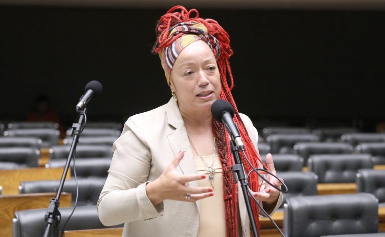 Deputada Daiana Santos fala ao microfone. Ela tem o cabelo vermelho, trançado e longo, usa um lenço colorido no cabelo e um blazer bege